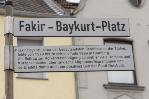 Straßenschild des Fakir-Baykurt-Platz, darunter ein Zusatzschild mit folgendem Text: Fakir Baykurt, einer der bedeutendsten Schriftsteller der Türkei, lebte von 1979 bis zu seinem Tode 1999 in Homberg. Als Beitrag zur Völkerverständigung schrieb er viele ROmane und Kurzgeschichten über türkische Migranten/ Migrantinnen und verbreitete damit auch ein positives Bild der Statd Duuisburg.