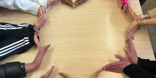 Auf dem Bild sind die Hände von 7 Personen zu sehen, die gemeinsam ein Herz auf einem Tisch formen.