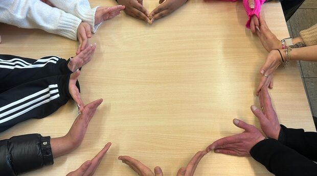 Auf dem Bild sind die Hände von 7 Personen zu sehen, die gemeinsam ein Herz auf einem Tisch formen.