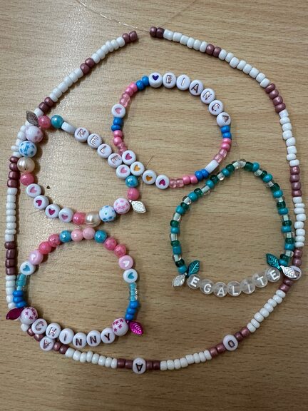 4 selbstgemachte Perlenarmbänder in den Farben weiß, rosa, blau und pink auf einem Tisch, umrandet werden die Armbänder von einer selbstegamchten Perlenkette